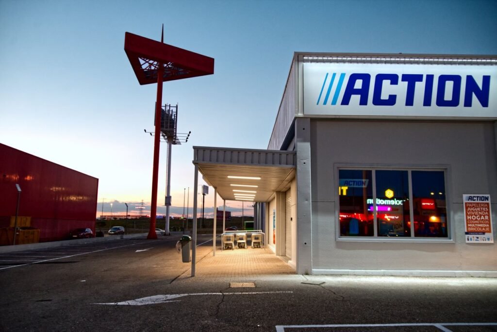 Fachada de loja com letreiro azul iluminado, vitrine de vidro e totem vermelho no estacionamento, ao entardecer