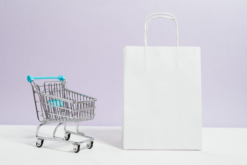 Sacola de papel branca com alças ao lado de um carrinho de compras em miniatura, sobre fundo lilás